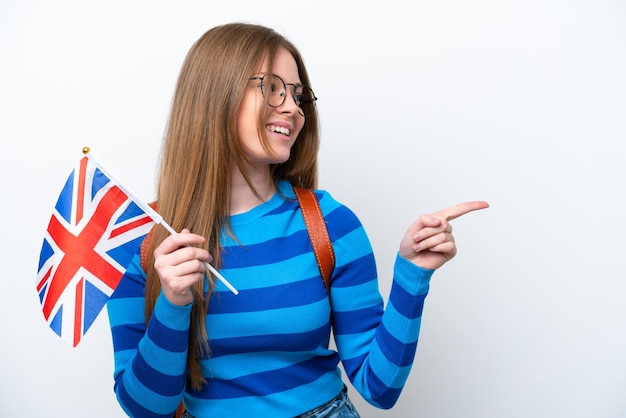흰색 배경에 격리된 영국 국기를 들고 옆으로 손가락을 가리키고 제품을 제시하는 젊은 백인 여성