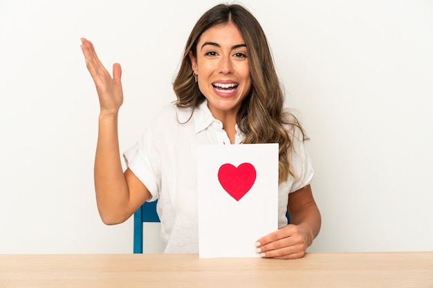 Young caucasian woman holding a valentines day card isolated receiving a pleasant surprise, excited and raising hands.