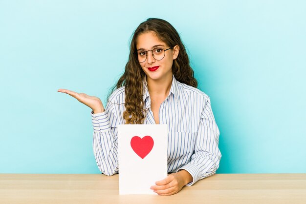 Giovane donna caucasica che tiene una carta di san valentino isolata mostrando uno spazio di copia su una palma e tenendo un'altra mano sulla vita.