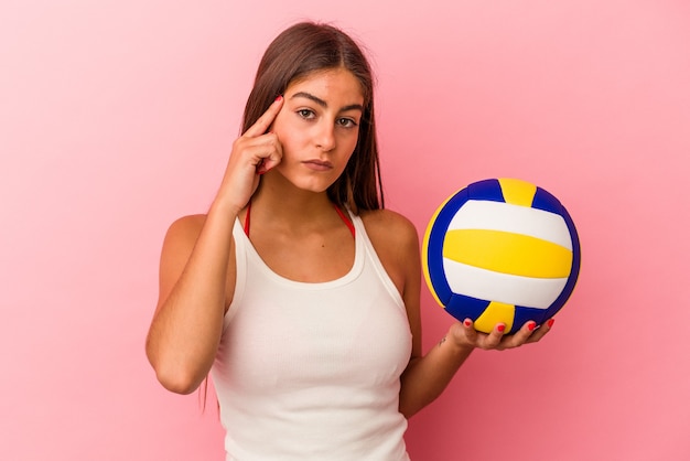 Young caucasian woman holding a volleyball ball isolated on pink background pointing temple with finger, thinking, focused on a task.