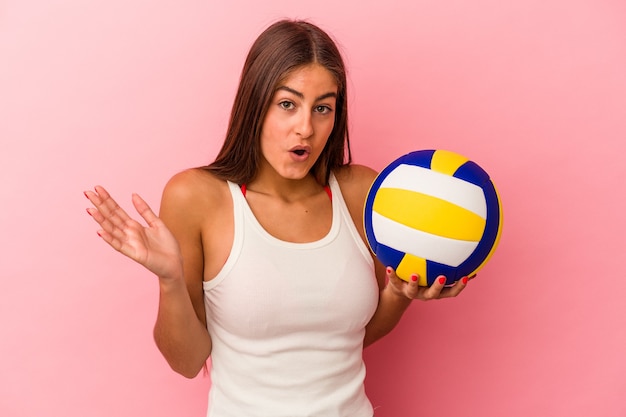 Young caucasian woman holding a volleyball ball isolated on pink background surprised and shocked.