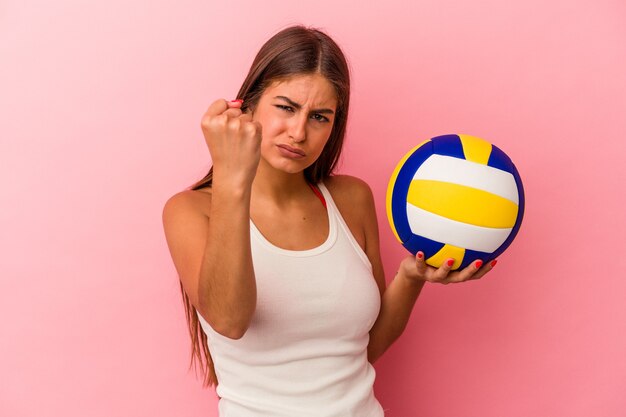 Young caucasian woman holding a volleyball ball isolated on pink wall showing fist, aggressive facial expression.