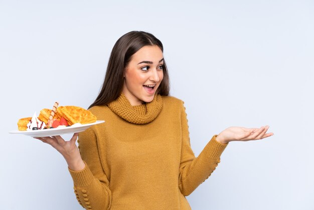 Young caucasian woman holding waffles isolated on blue wall with surprise facial expression