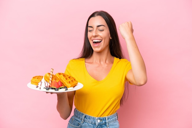 Young caucasian woman holding waffles isolated on pink background celebrating a victory