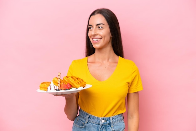 Young caucasian woman holding waffles isolated on pink background looking side