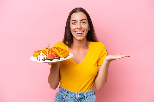 Photo young caucasian woman holding waffles isolated on pink background with shocked facial expression