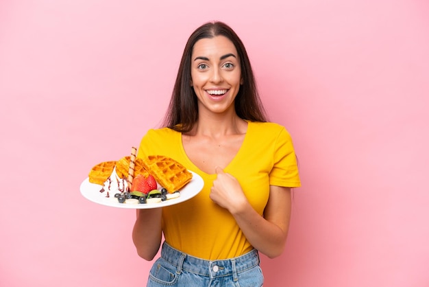 Young caucasian woman holding waffles isolated on pink background with surprise facial expression