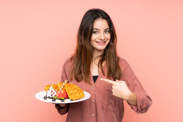 Young caucasian woman holding waffles isolated on pink wall with surprise facial expression