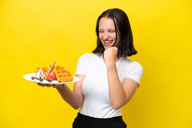 Young caucasian woman holding waffles isolated on yellow background celebrating a victory