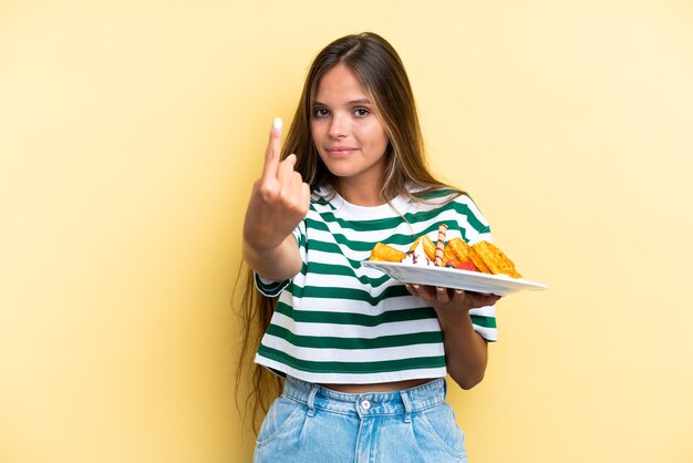 Young caucasian woman holding waffles isolated on yellow background doing coming gesture