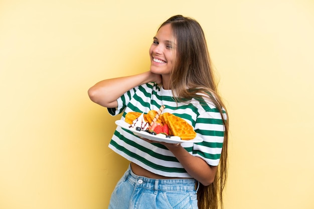 Young caucasian woman holding waffles isolated on yellow background laughing