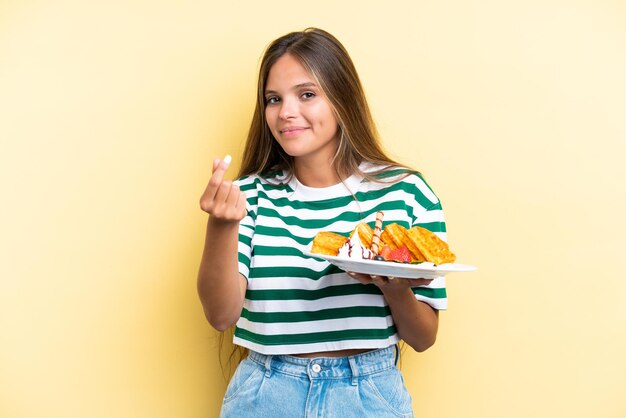 Young caucasian woman holding waffles isolated on yellow background making money gesture