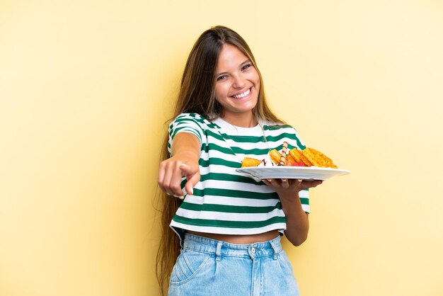 Young caucasian woman holding waffles isolated on yellow background pointing front with happy expression