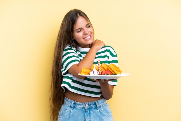 Young caucasian woman holding waffles isolated on yellow background suffering from pain in shoulder for having made an effort