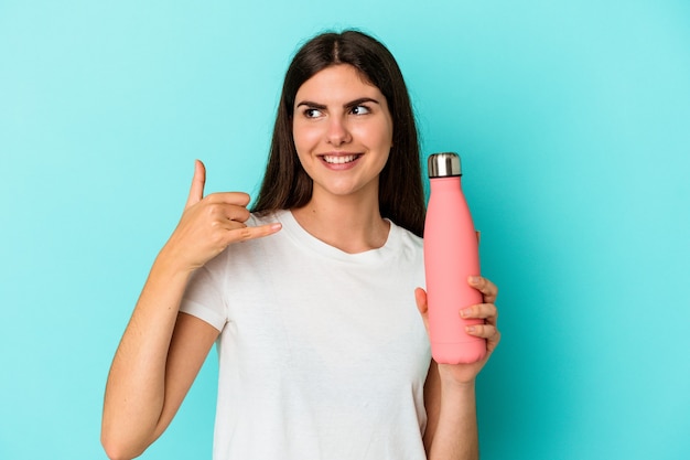 Young caucasian woman holding water bottle isolated on blue background showing a mobile phone call gesture with fingers.