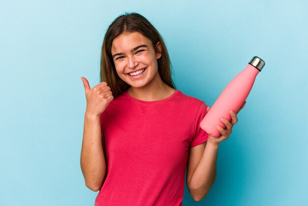 Giovane donna caucasica in possesso di una bottiglia d'acqua isolata su sfondo blu sorridente e alzando il pollice thumb