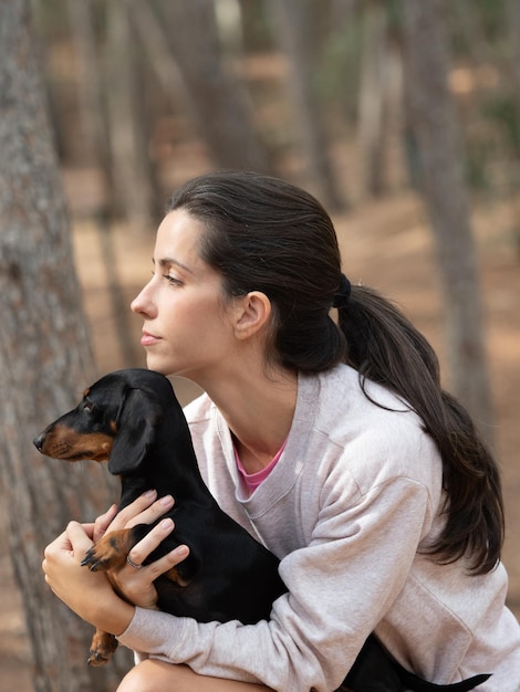 Young caucasian woman hugging black smooth haired miniature\
dachshund in forest