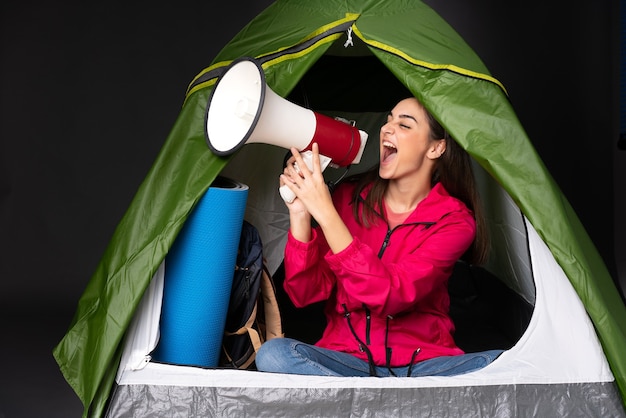 Young caucasian woman inside a camping green tent shouting through a megaphone