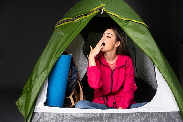 Young caucasian woman inside a camping green tent yawning and covering wide open mouth with hand