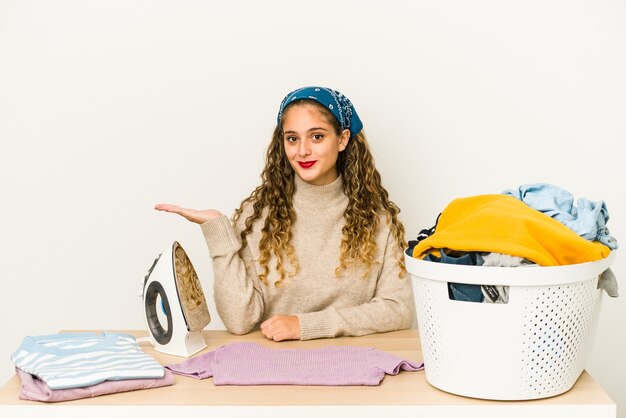 Young caucasian woman ironing clothes isolated showing a copy space on a palm and holding another hand on waist.