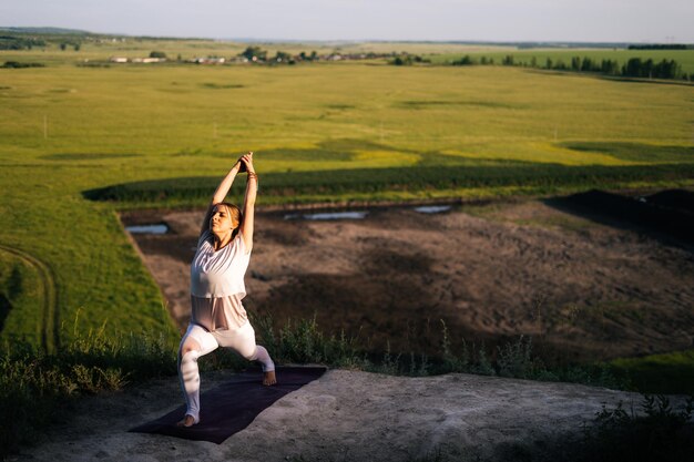 Young Caucasian woman is performing warrior yoga pose on top of rock background of flowing river