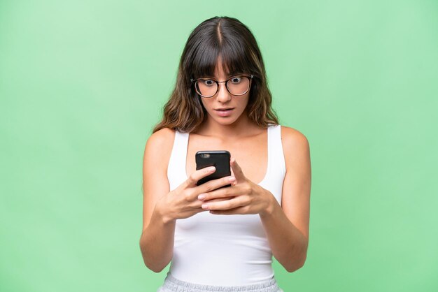 Young caucasian woman over isolated background looking at the\
camera while using the mobile with surprised expression