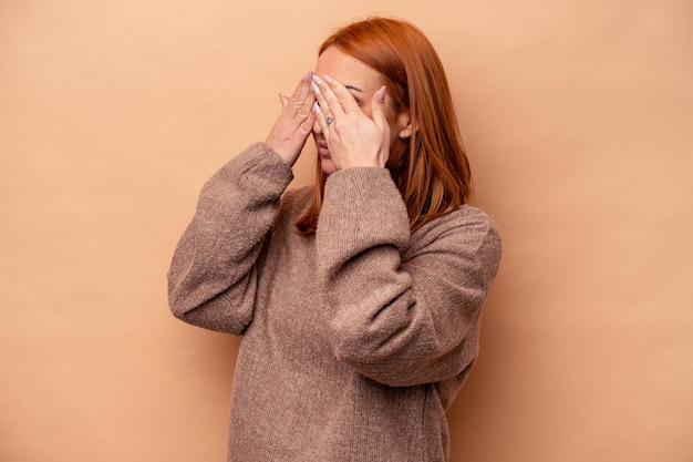 Giovane donna caucasica isolata su sfondo beige che copre gli occhi impauriti con le mani.