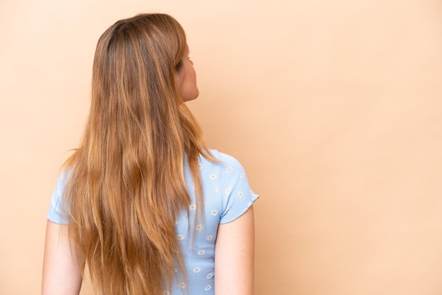 Giovane donna caucasica isolata su sfondo beige in posizione posteriore e guardando indietro