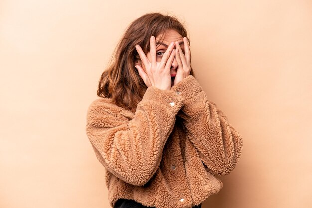 Young caucasian woman isolated on beige background blink through fingers frightened and nervous