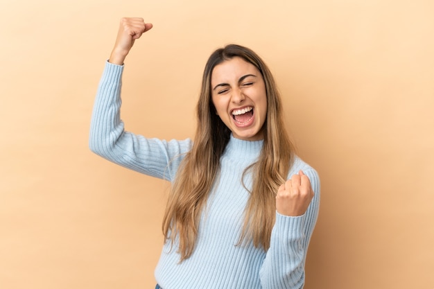 Giovane donna caucasica isolata su fondo beige che celebra una vittoria