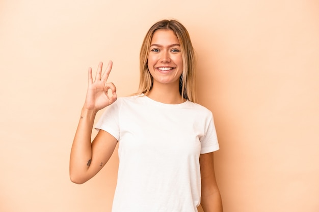 Young caucasian woman isolated on beige background cheerful and confident showing ok gesture.