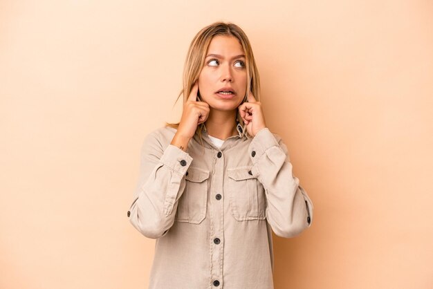 Young caucasian woman isolated on beige background covering ears with fingers, stressed and desperate by a loudly ambient.