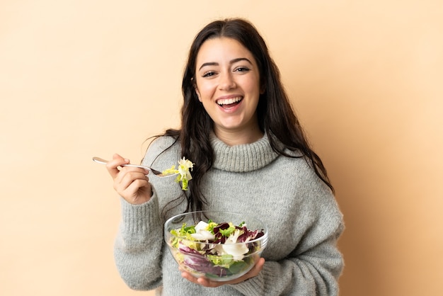 Young caucasian woman isolated on beige background holding a bowl of salad with happy expression