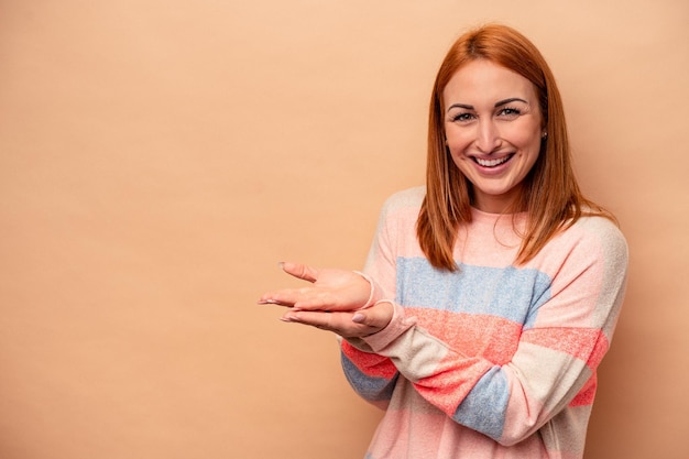 Young caucasian woman isolated on beige background holding a copy space on a palm.