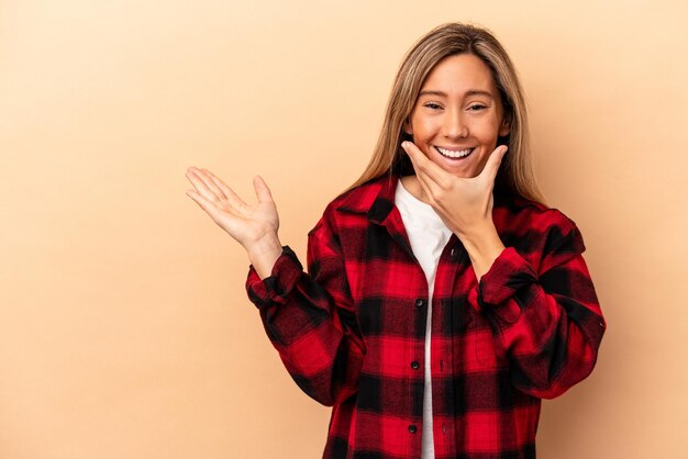 Young caucasian woman isolated on beige background holds copy space on a palm, keep hand over cheek. Amazed and delighted.