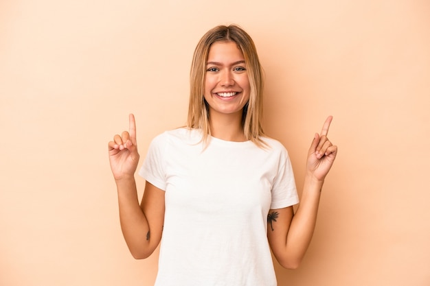 Young caucasian woman isolated on beige background indicates with both fore fingers up showing a blank space.