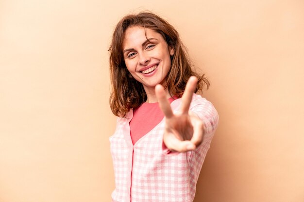 Young caucasian woman isolated on beige background joyful and carefree showing a peace symbol with fingers