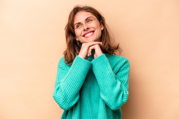 Young caucasian woman isolated on beige background keeps hands under chin is looking happily aside