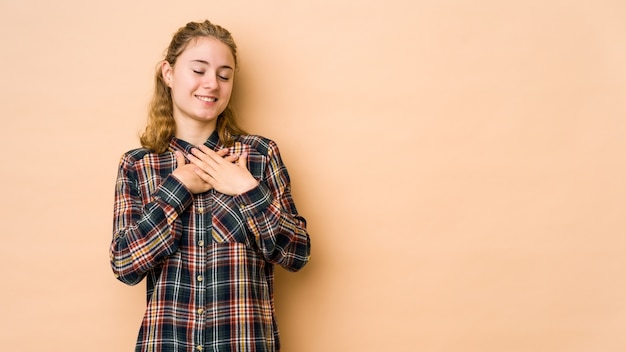 ベージュの背景に孤立した若い白人女性は、心に手を置いて笑って、幸せの概念。