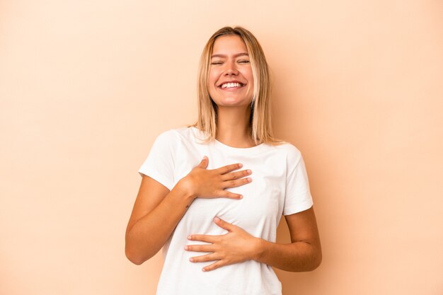 Young caucasian woman isolated on beige background laughs happily and has fun keeping hands on stomach.