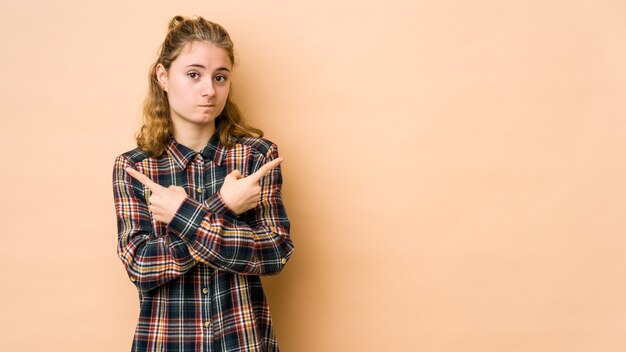 Photo young caucasian woman isolated on beige background points sideways, is trying to choose between two options.