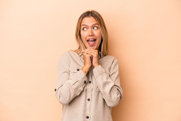 Young caucasian woman isolated on beige background praying for luck, amazed and opening mouth looking to front.