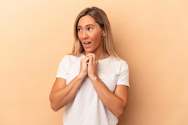 Giovane donna caucasica isolata su fondo beige pregando per la fortuna, stupita e aprendo la bocca guardando in avanti.