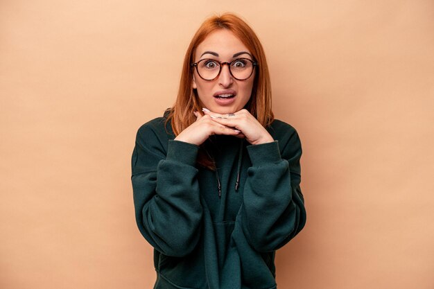 Young caucasian woman isolated on beige background praying for luck, amazed and opening mouth looking to front.