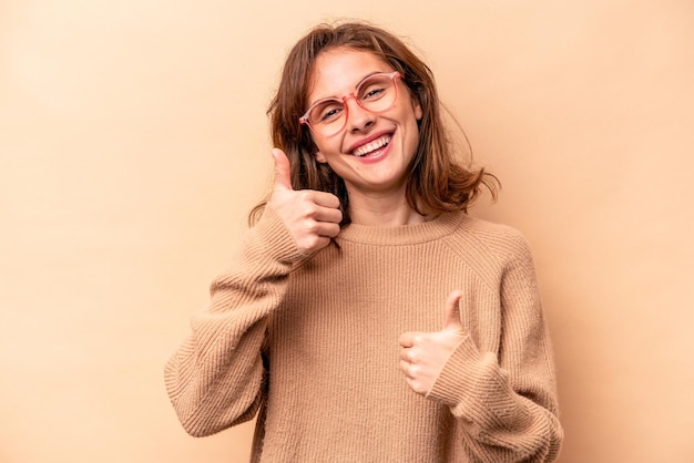 ベージュの背景に分離された若い白人女性は、笑顔と自信を持って両方の親指を上げます