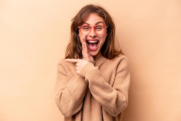 Young caucasian woman isolated on beige background saying a gossip pointing to side reporting something