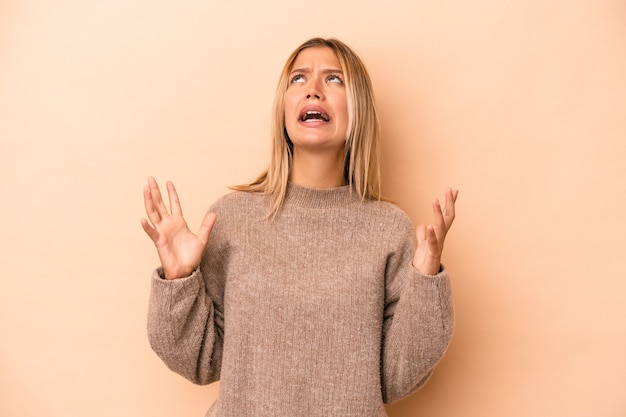 Young caucasian woman isolated on beige background screaming to the sky, looking up, frustrated.