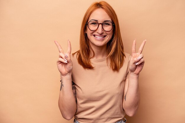 Young caucasian woman isolated on beige background showing victory sign and smiling broadly.