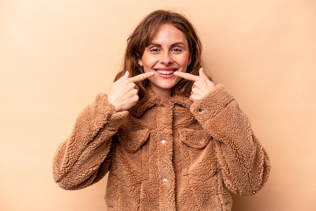 Young caucasian woman isolated on beige background smiles pointing fingers at mouth