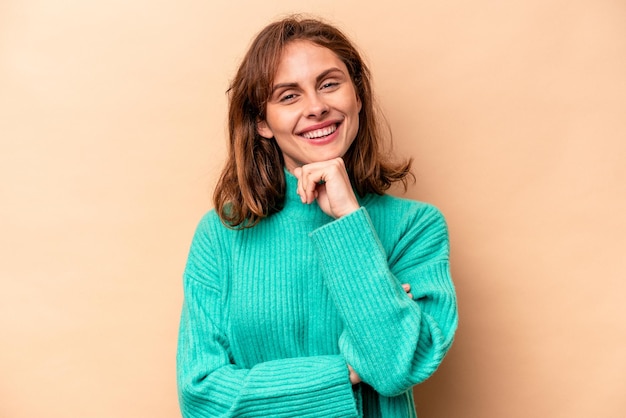 Young caucasian woman isolated on beige background smiling happy and confident touching chin with hand